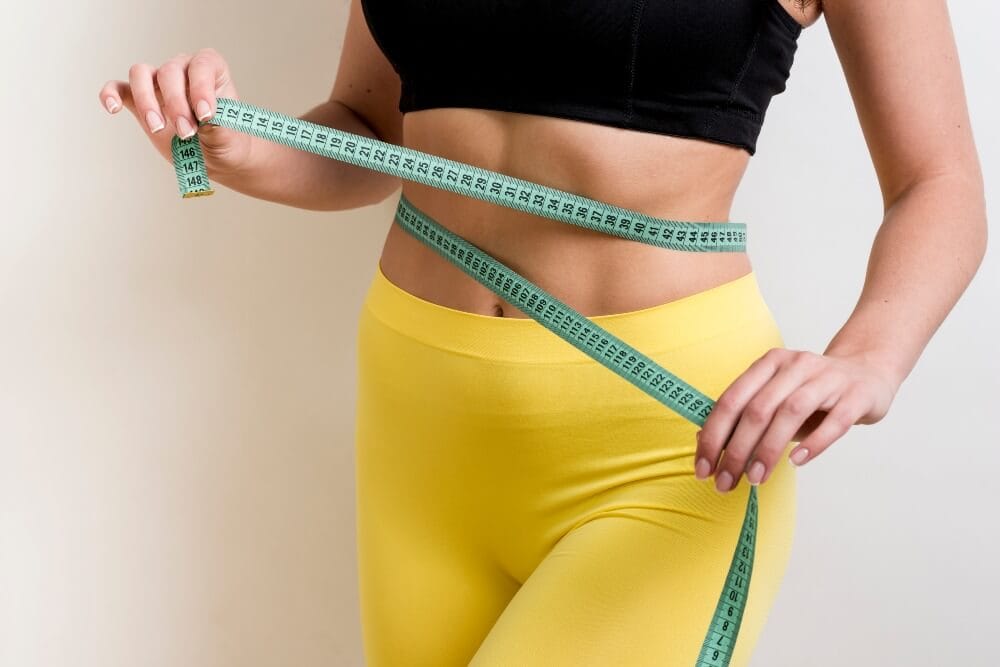 Woman in yellow leggings measuring her waist with a measuring tape