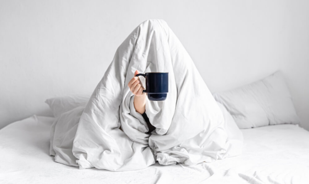 Woman wrapped in a white blanket on a bed holding a cup of coffee