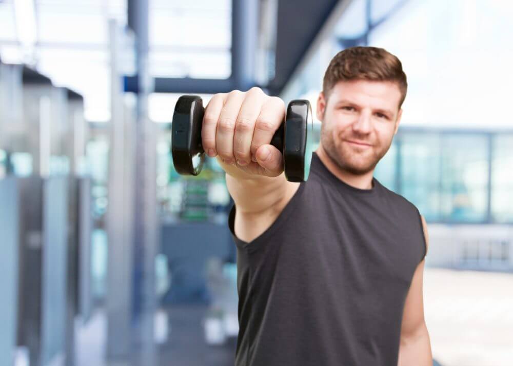 Smiling man doing a dumbbell front raise in a bright gym