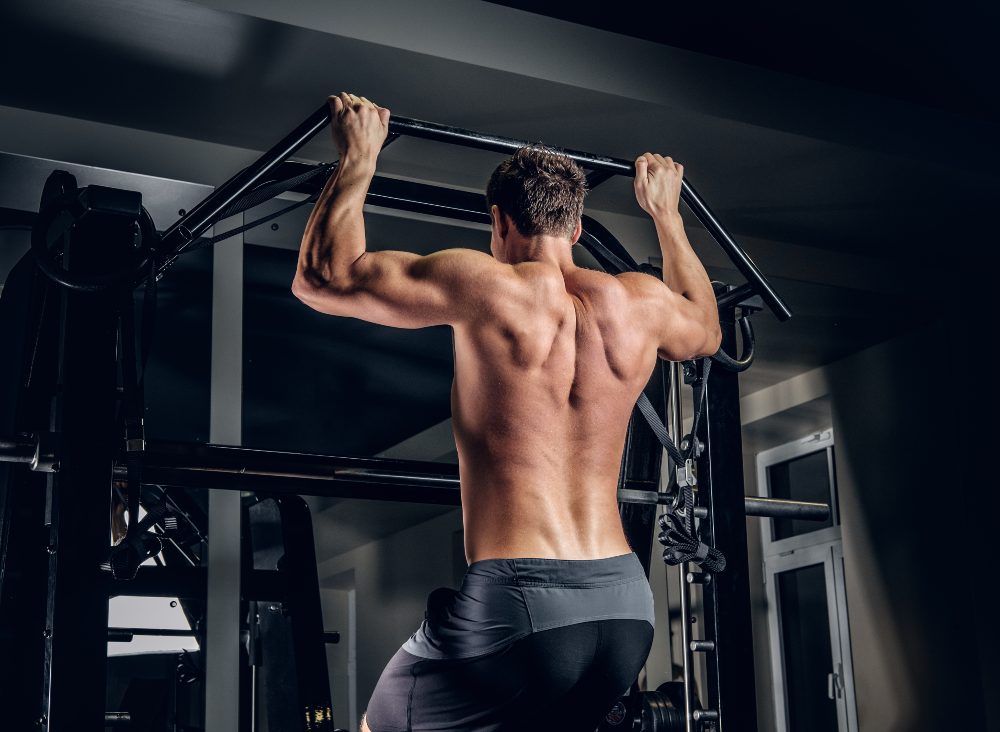 Shirtless man doing pull ups on a bar in a dark gym, seen from behind