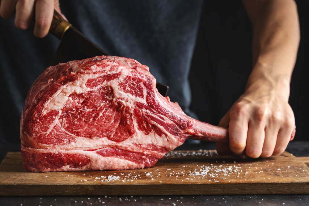 Hands cutting a large raw beef rib cut on a wooden board