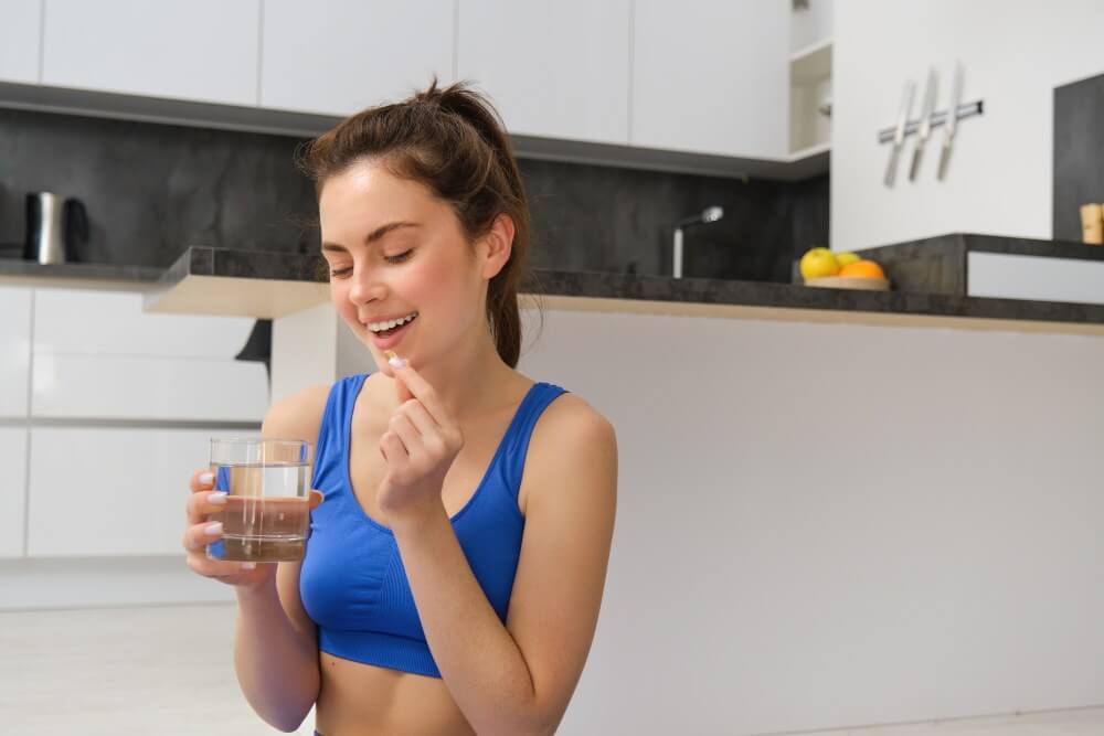 Smiling woman in sportswear taking a vitamin with a glass of water at home