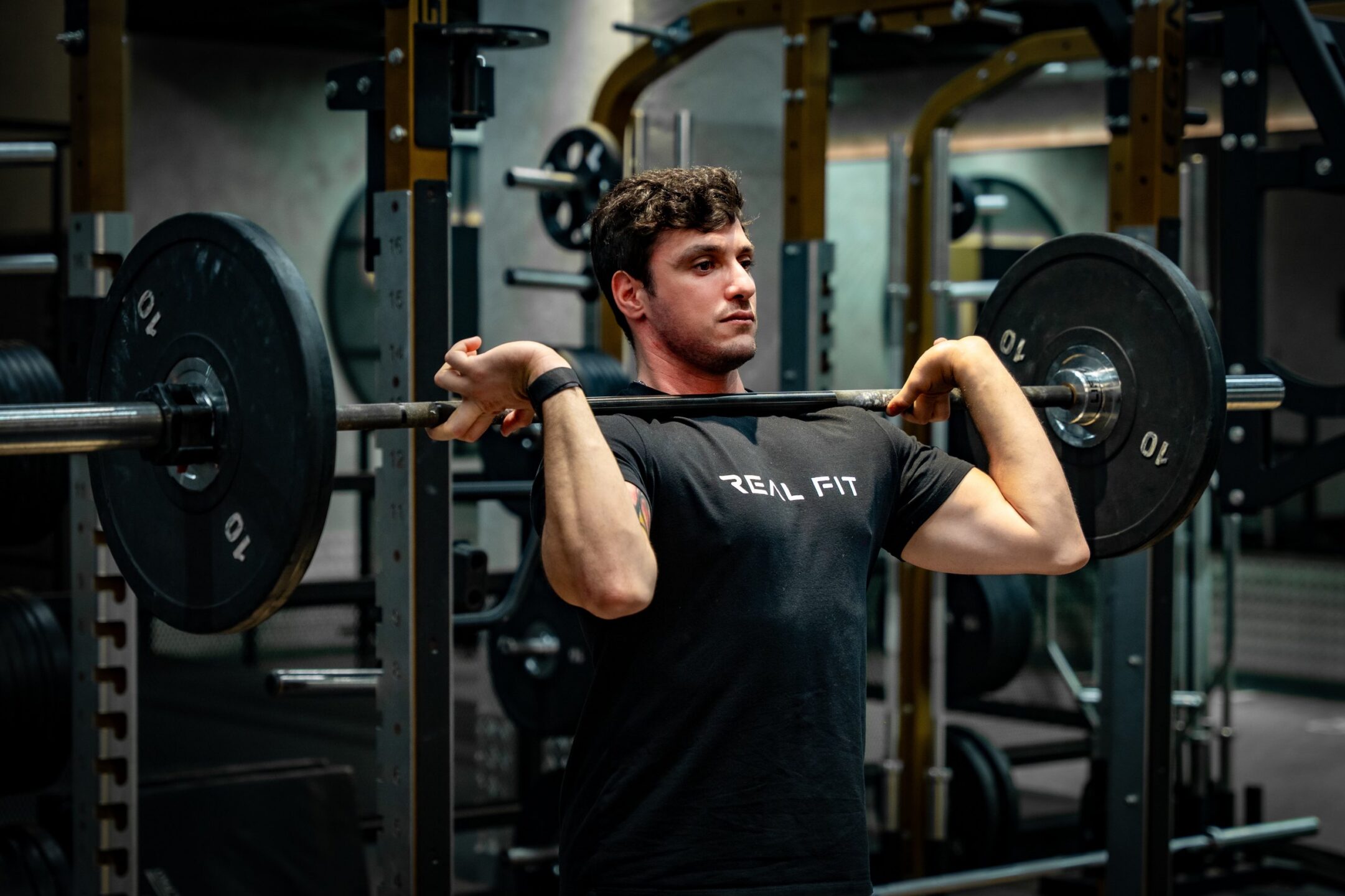 Man in a Real Fit T-shirt holding a loaded barbell at shoulder height in the gym