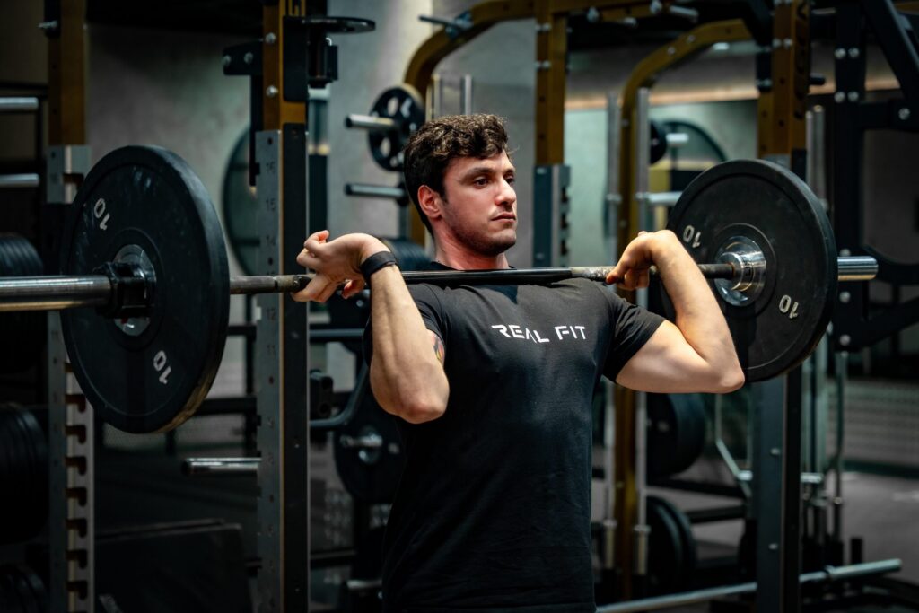 Marcello, Real Fit personal trainer, holding a loaded barbell at shoulder height in the gym