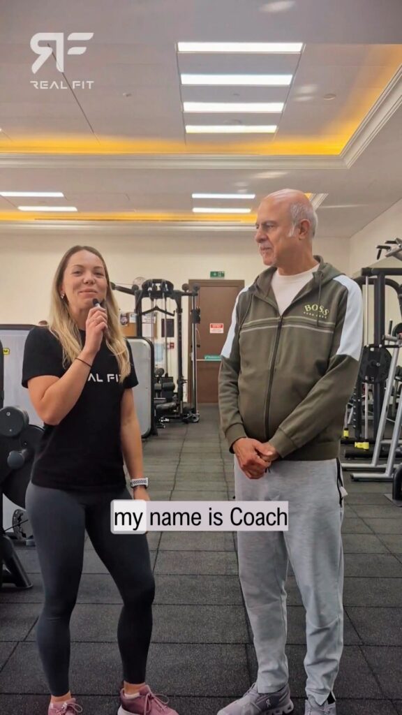 Coach Lucy with a microphone introducing client Basam in the gym